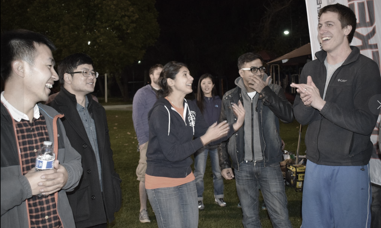 A group of young coworkers laughing and clapping together on a lawn at night