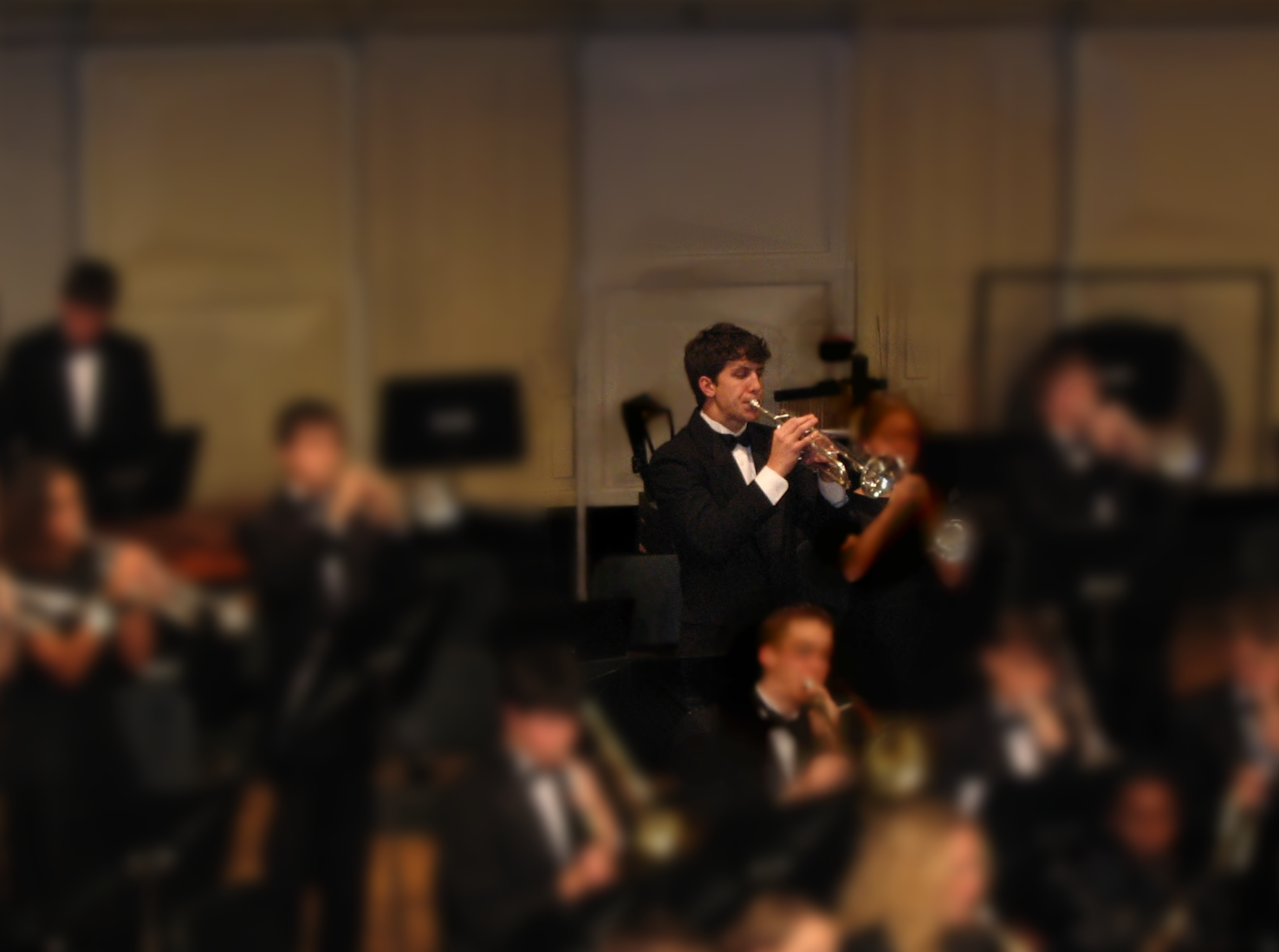 Young man in a tuxedo playing trumpet in a concert band, surrounded by blurred musicians