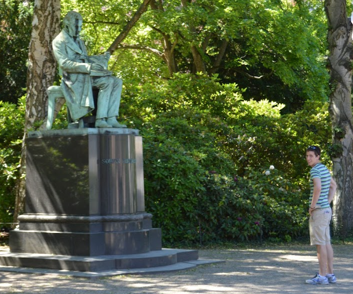 Young man in a striped shirt looking back beside a bronze statue of Søren Kierkegaard seated in a park