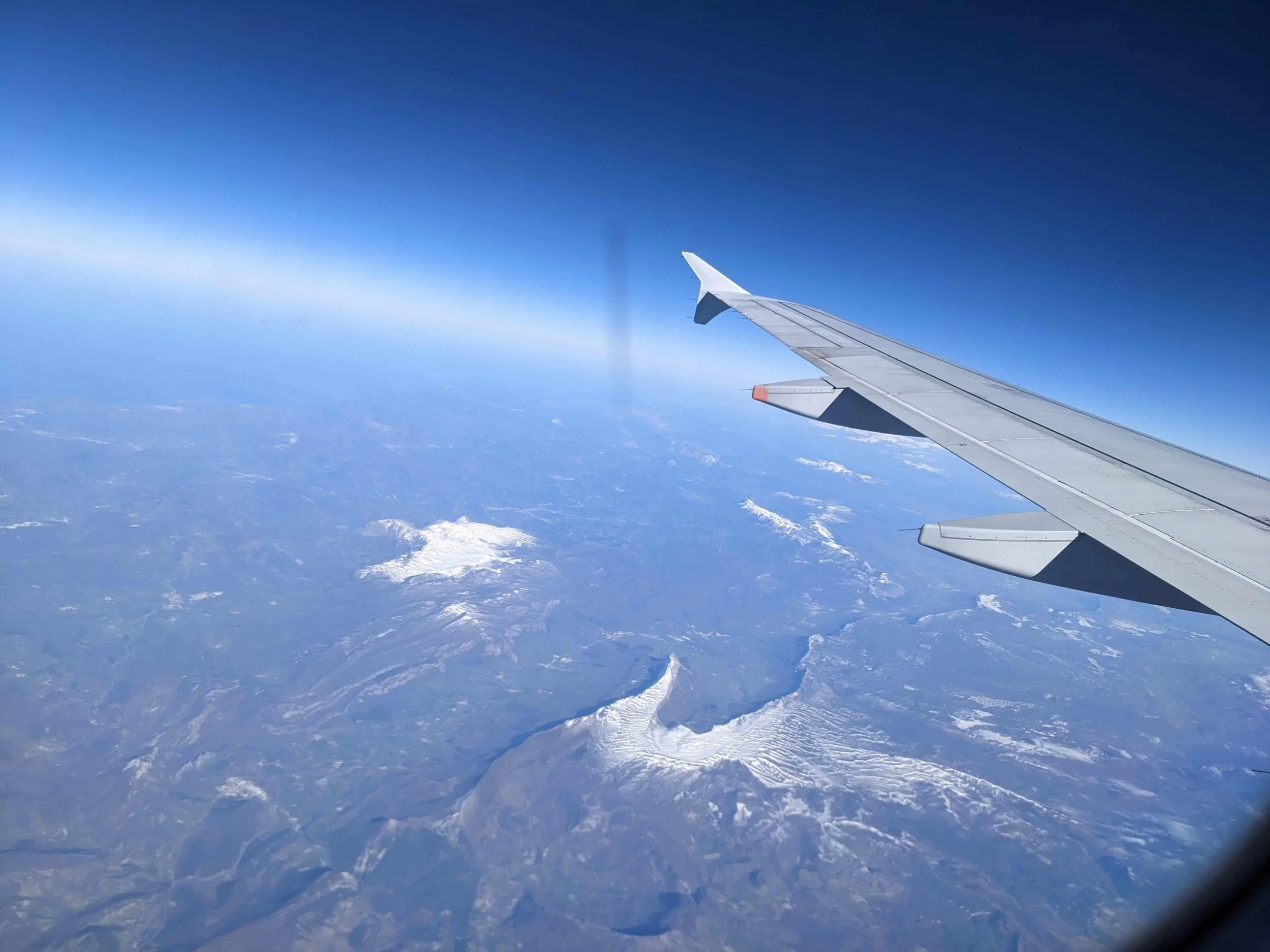 View from a plane window of the wing over snow-dusted mountains under a deep blue sky