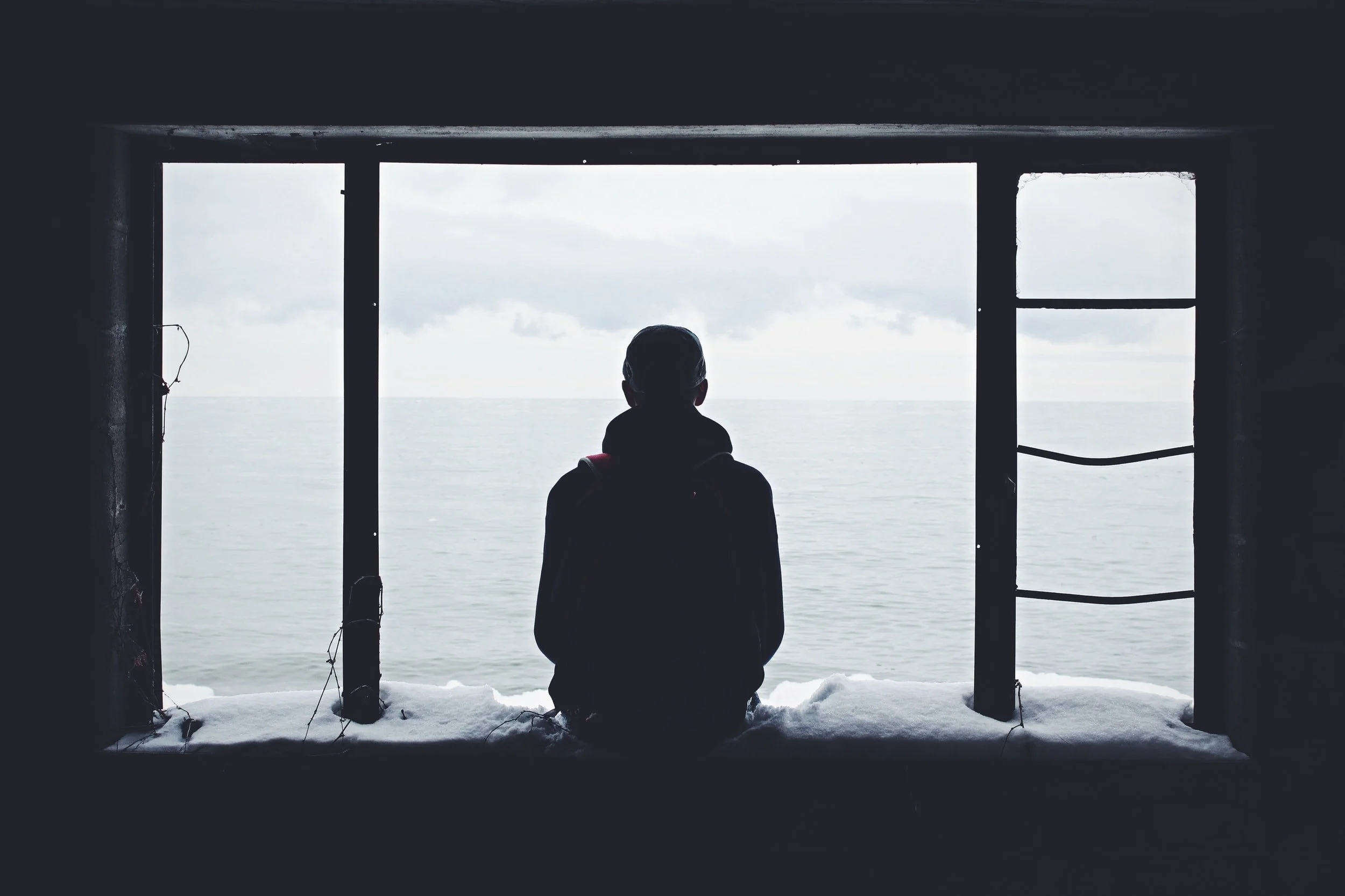 Silhouette of a person sitting on a snowy window ledge, looking out at a gray sea