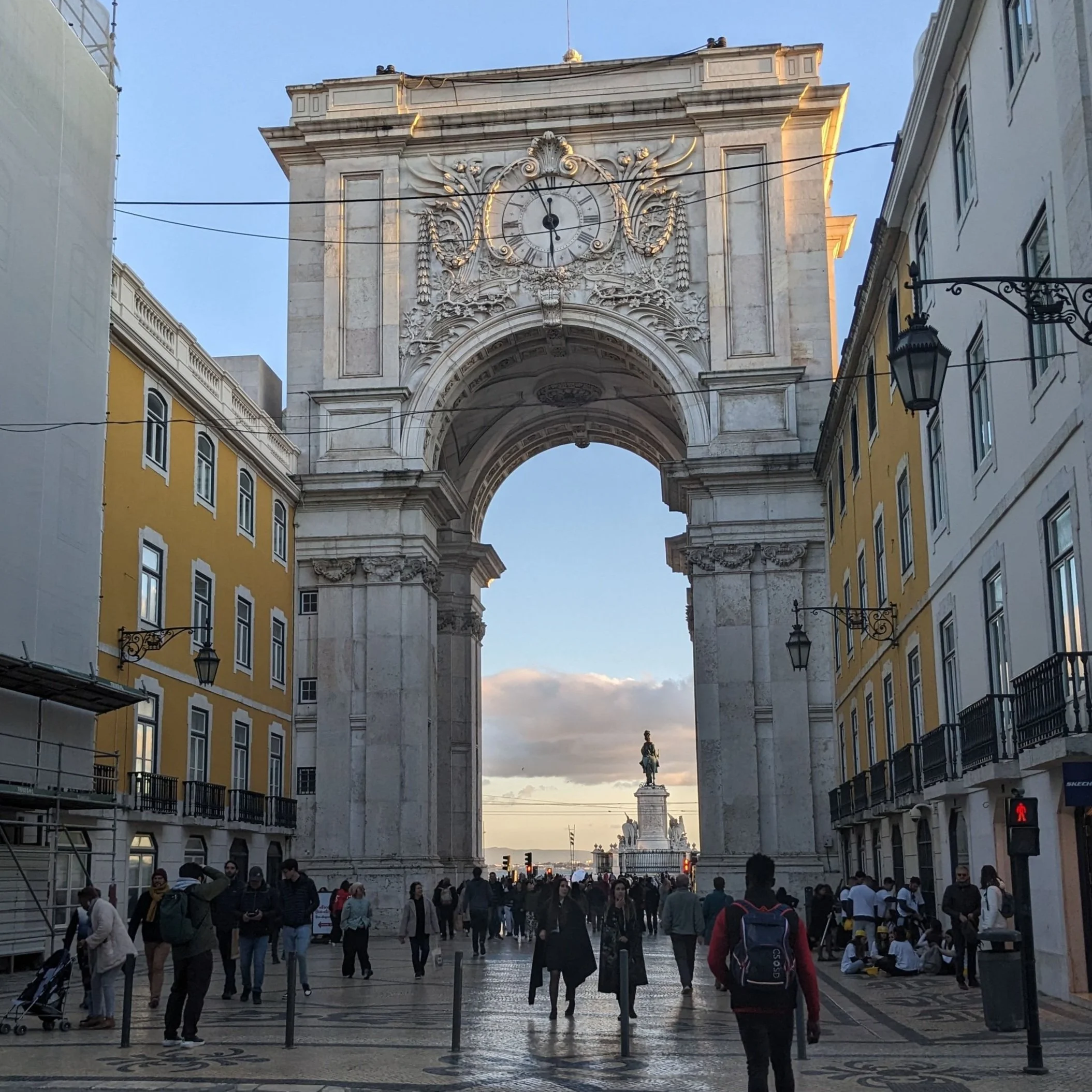 Large stone arch with a clock over a busy pedestrian street in Lisbon, a statue visible through it