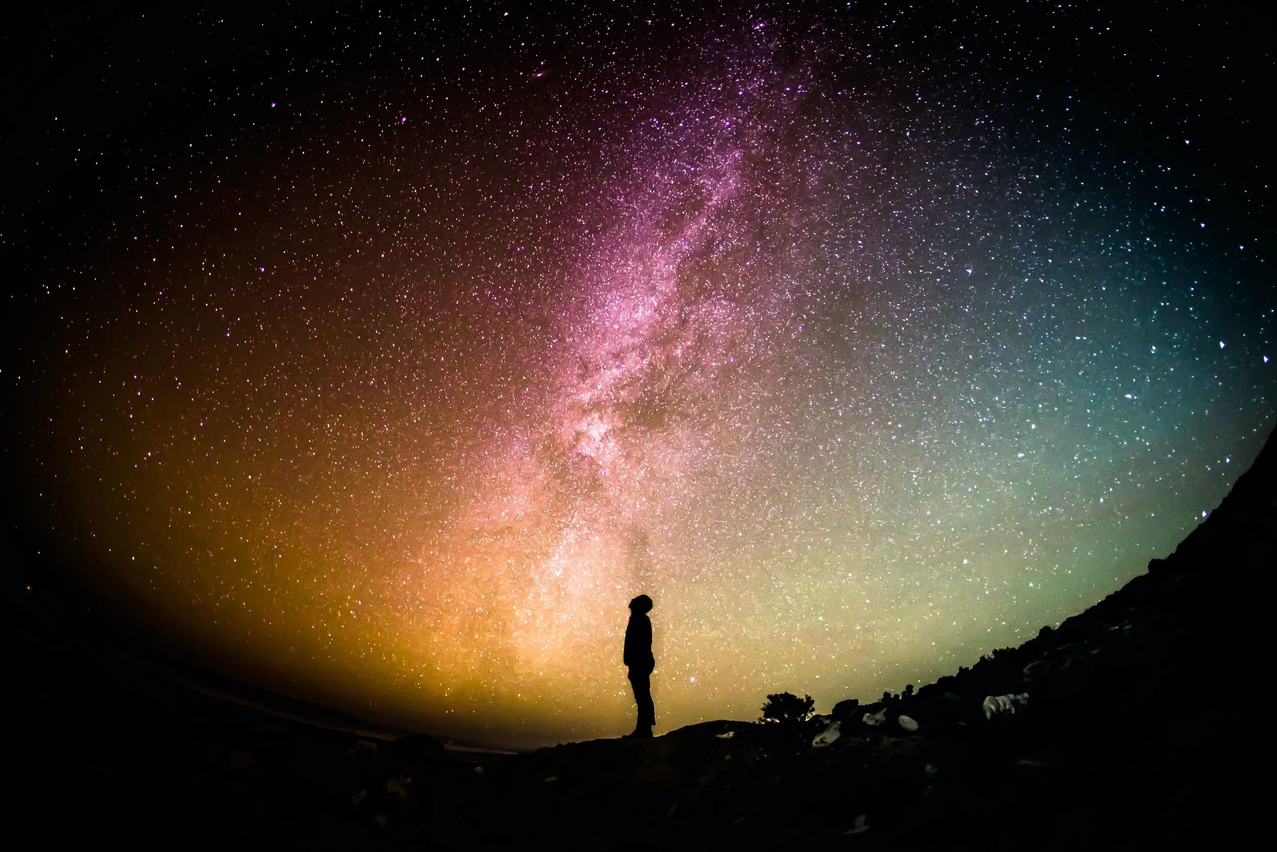Silhouette of a person standing on a hill looking up at a colorful, star-filled night sky and the Milky Way