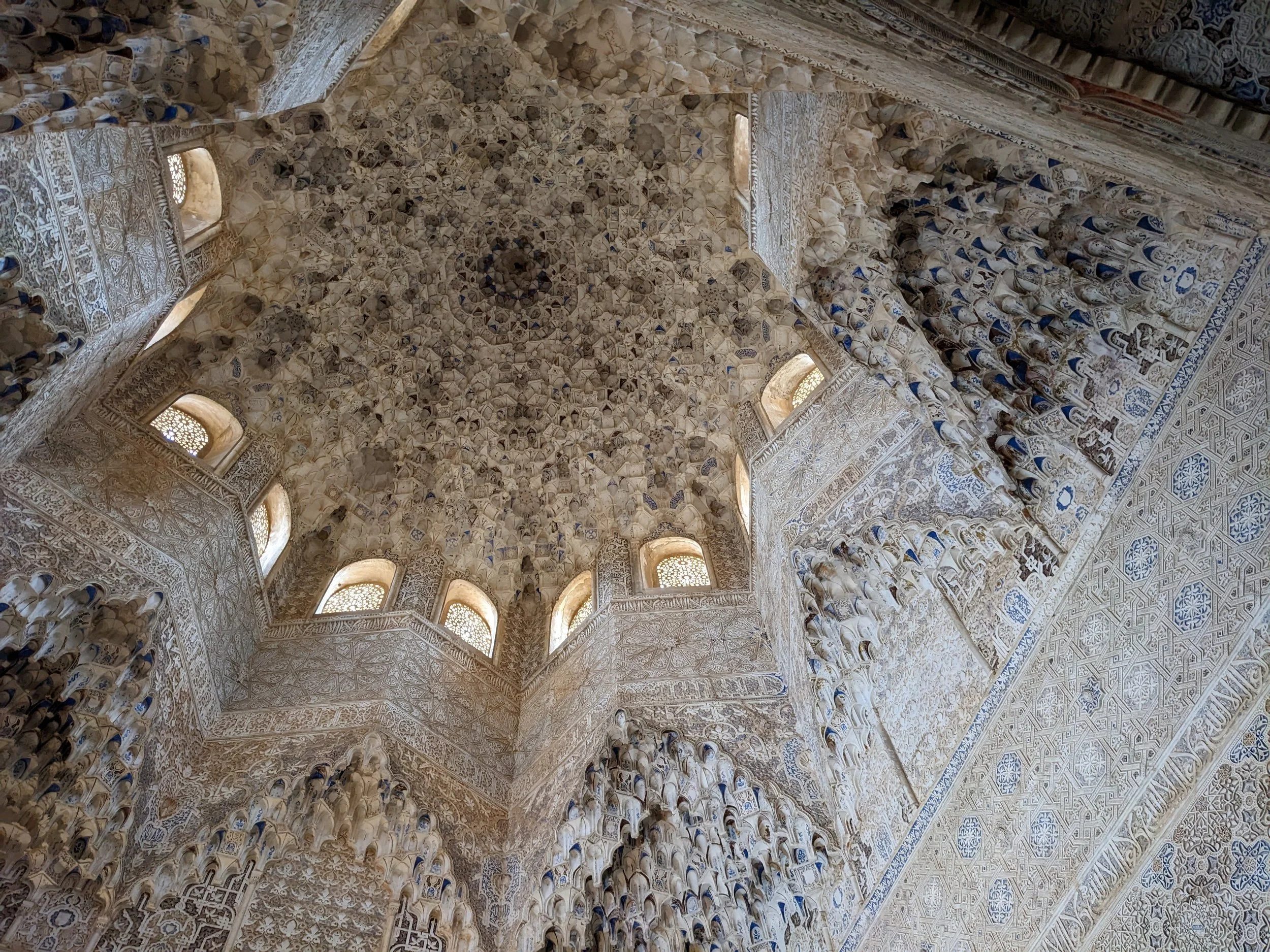 Looking up at an intricately carved stone dome with small arched windows inside the Alhambra