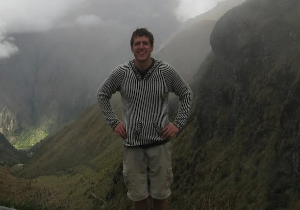 Smiling man in a striped sweater standing on a misty mountain pass in Peru, with green valleys behind him
