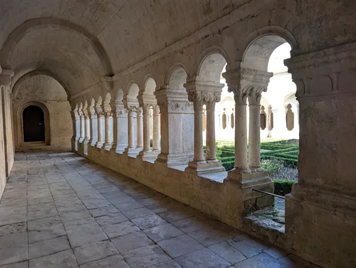 Vaulted stone cloister walkway with carved columns and arches opening onto a hedged garden