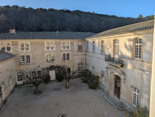 Courtyard of old stone abbey buildings with tile roofs and bare trees below a wooded hill