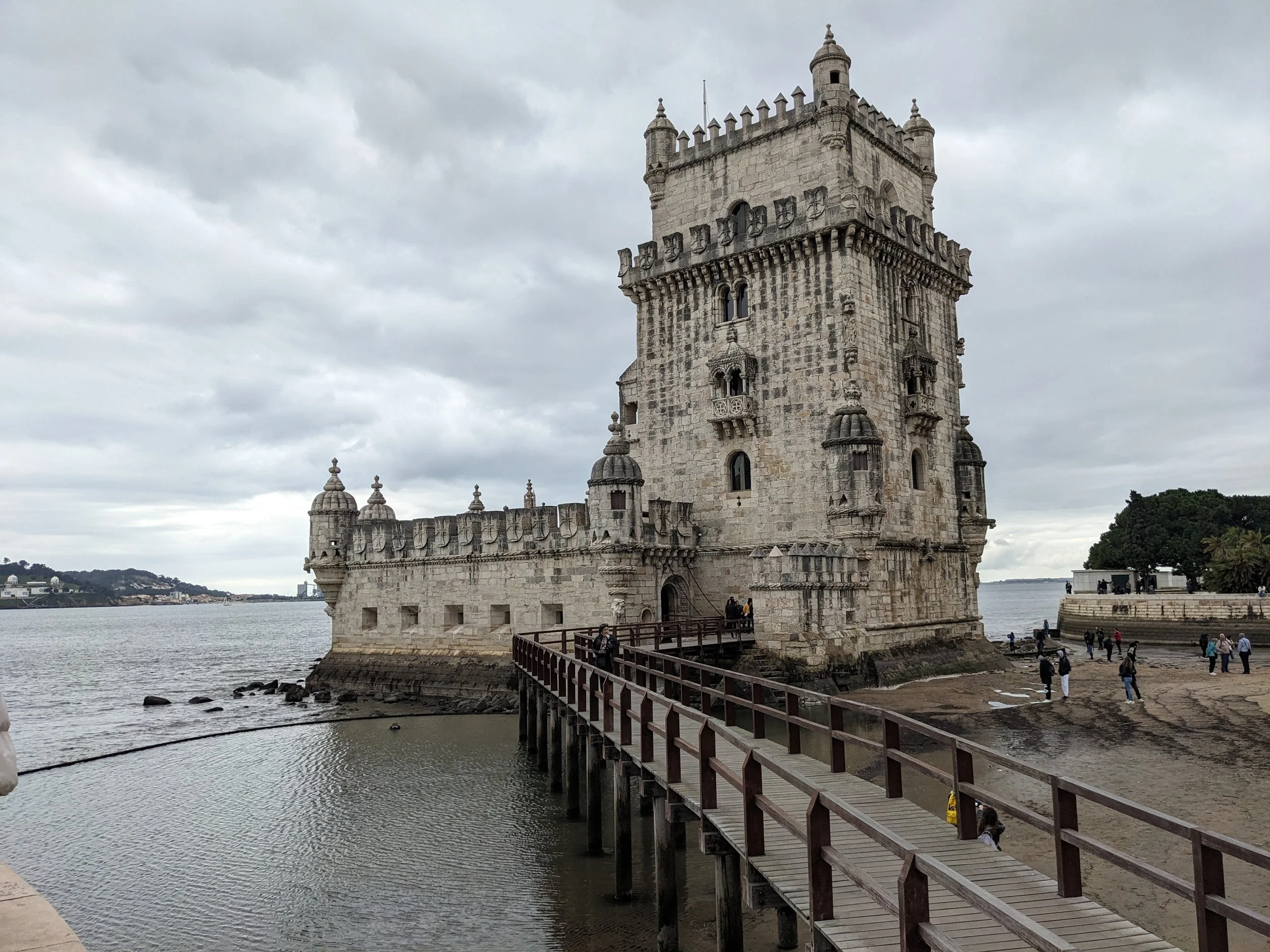 Belem Tower, an ornate stone fortress on the water, reached by a wooden footbridge under cloudy skies