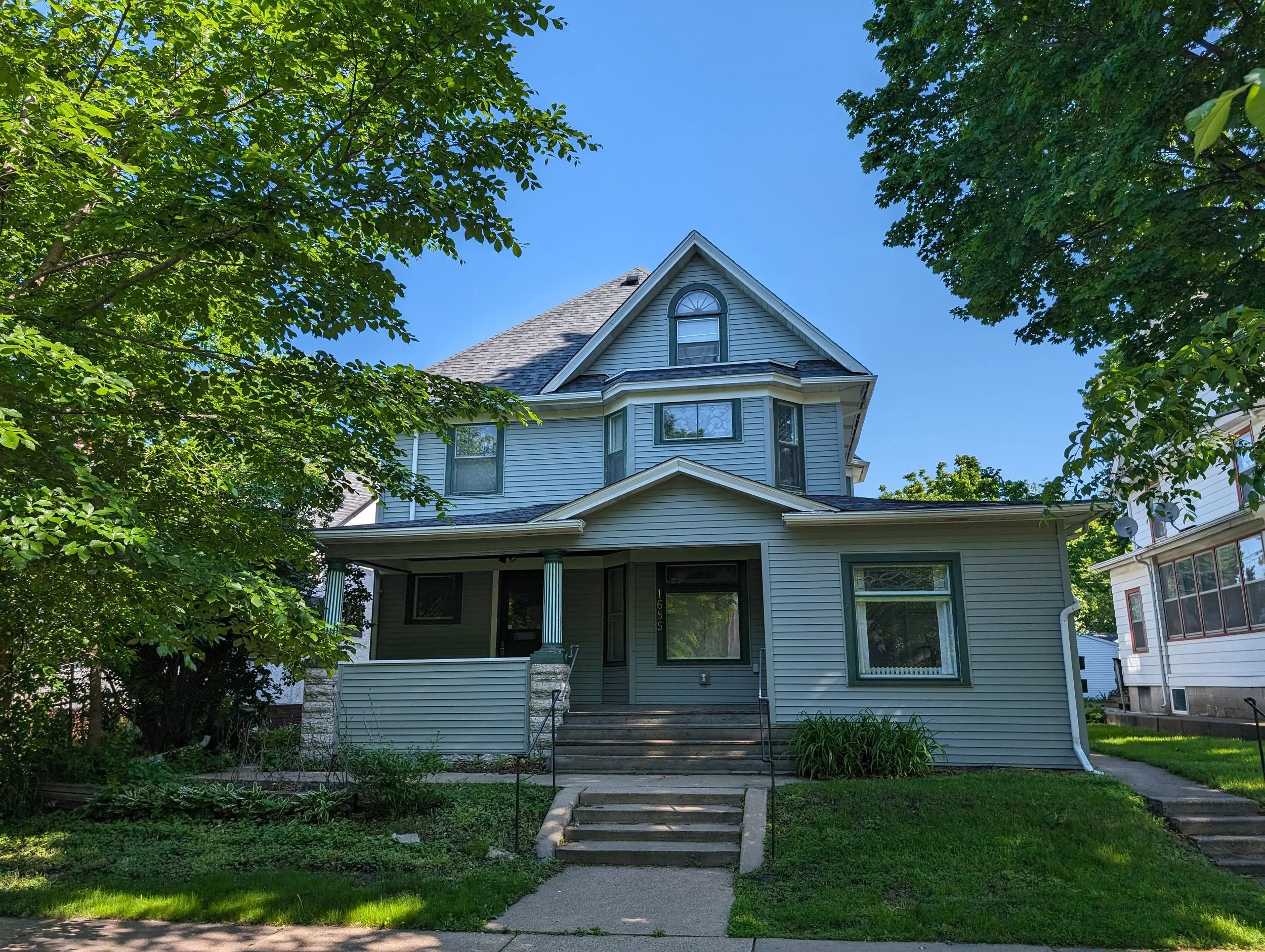 Old two-story gray-green house with a front porch, framed by leafy trees under a blue sky