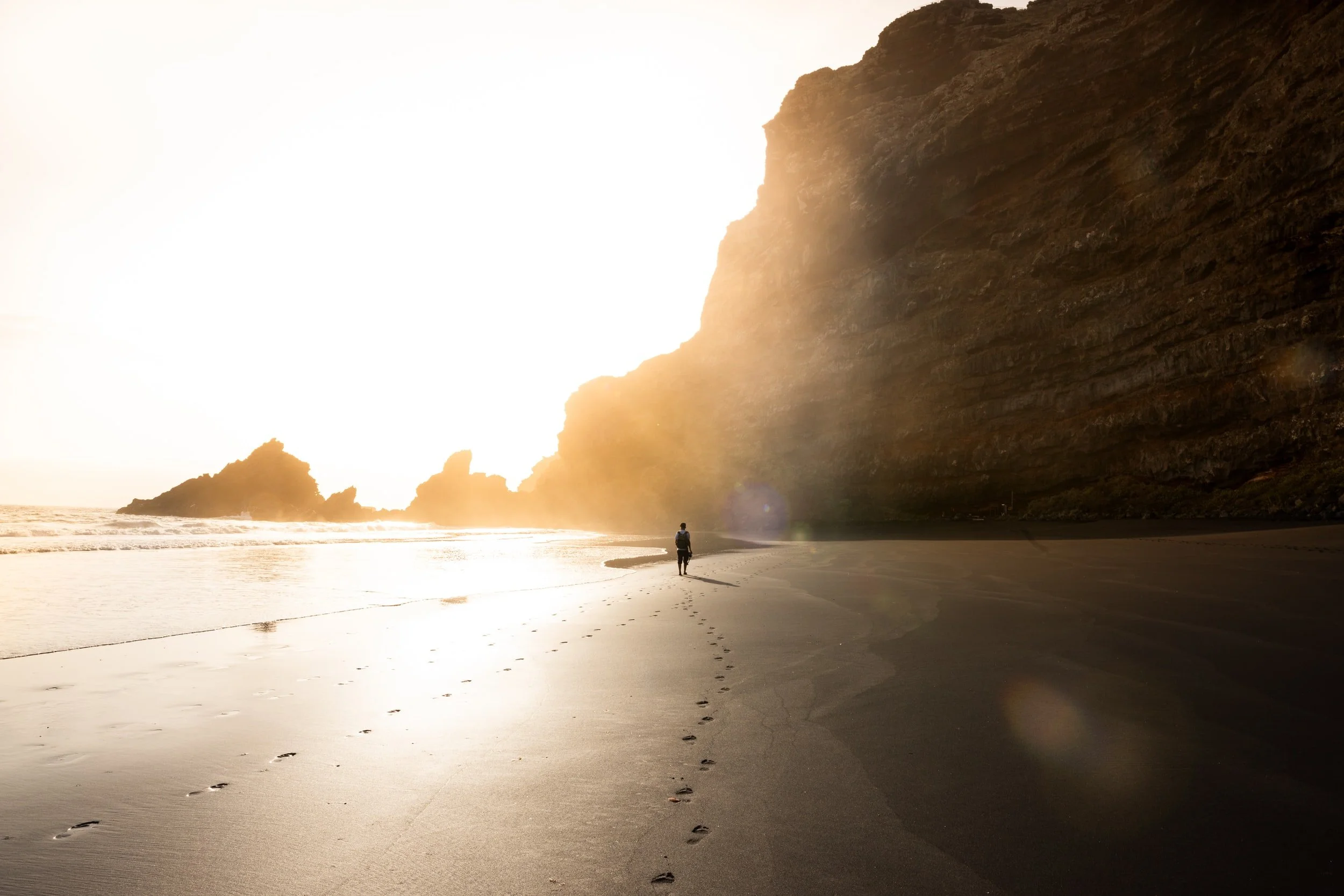 A lone person walks along a dark sand beach beneath a tall cliff, sunlight glowing over the ocean