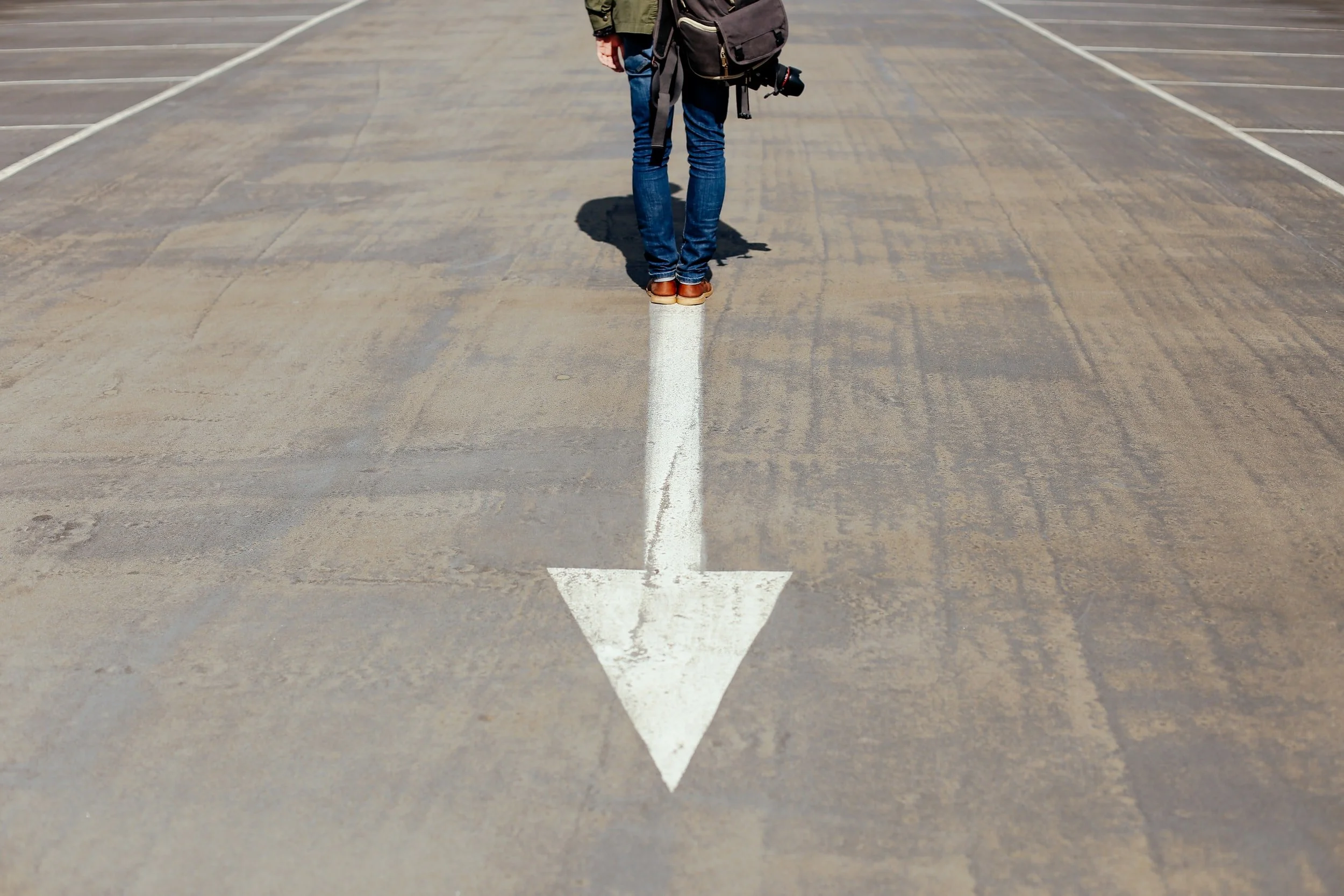 A person with a camera bag stands at the tail of a white arrow painted on an empty parking lot