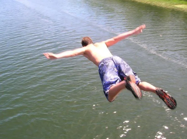 A shirtless young man in swim trunks leaping arms out toward a lake
