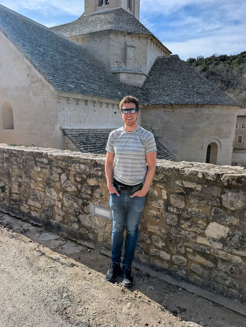 Smiling man in sunglasses and a striped shirt standing by a stone wall in front of the abbey church
