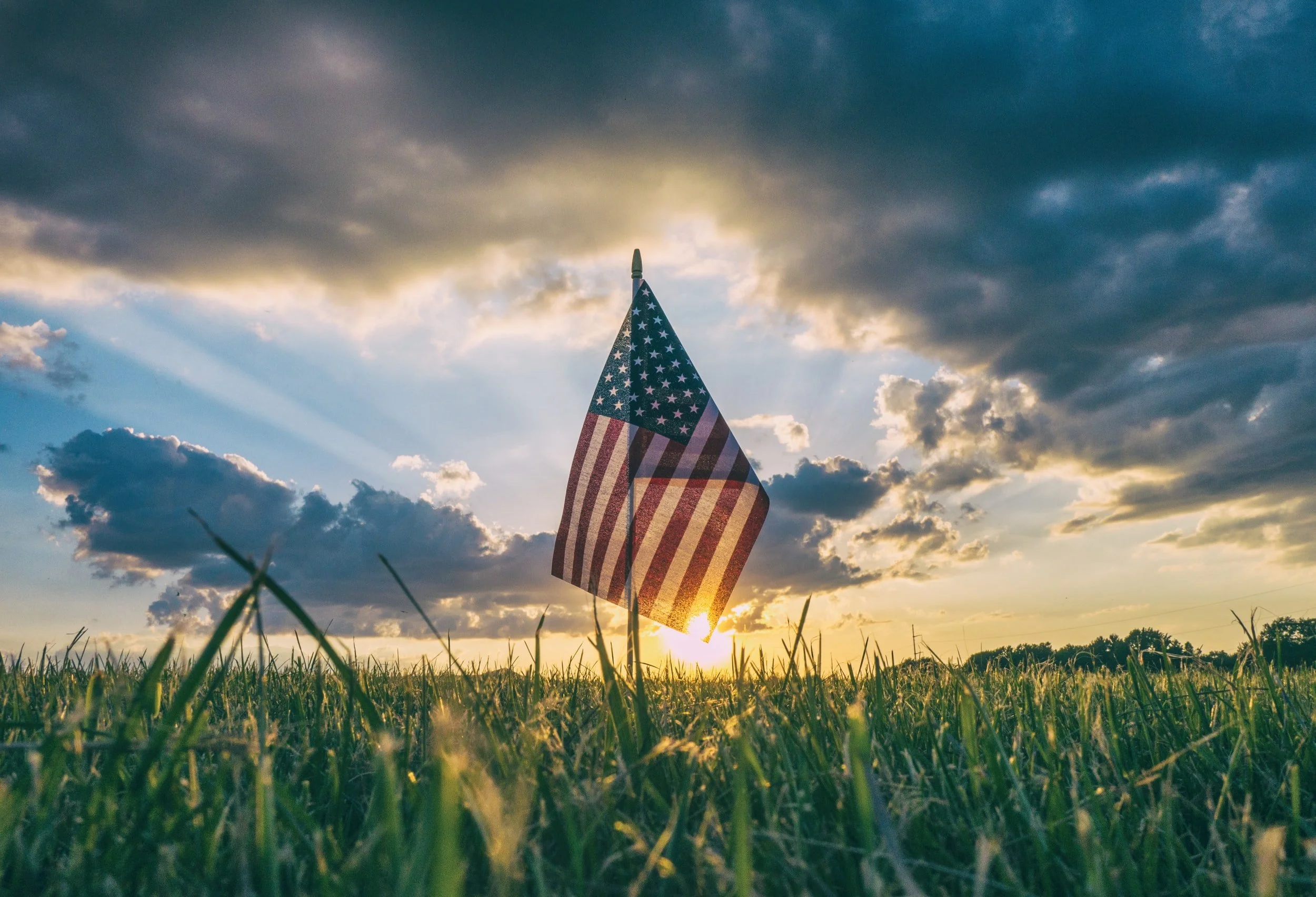 A small American flag in tall grass, backlit by the setting sun under dark clouds