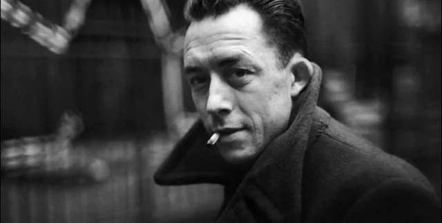 Black and white photo of Albert Camus in a heavy coat with a cigarette in his mouth