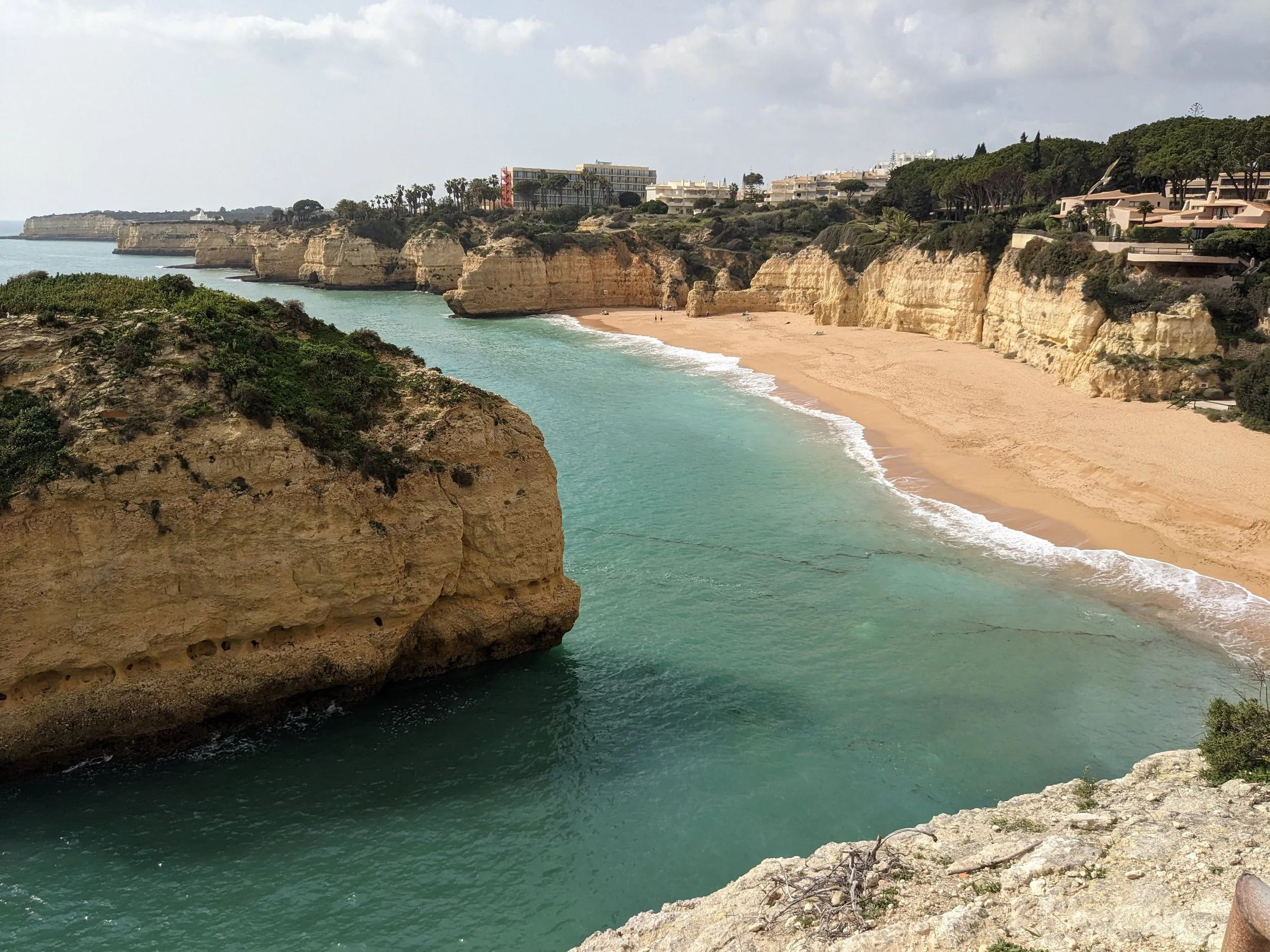 Sandy cove framed by tall yellow cliffs and green water on the Algarve coast, with hotels above