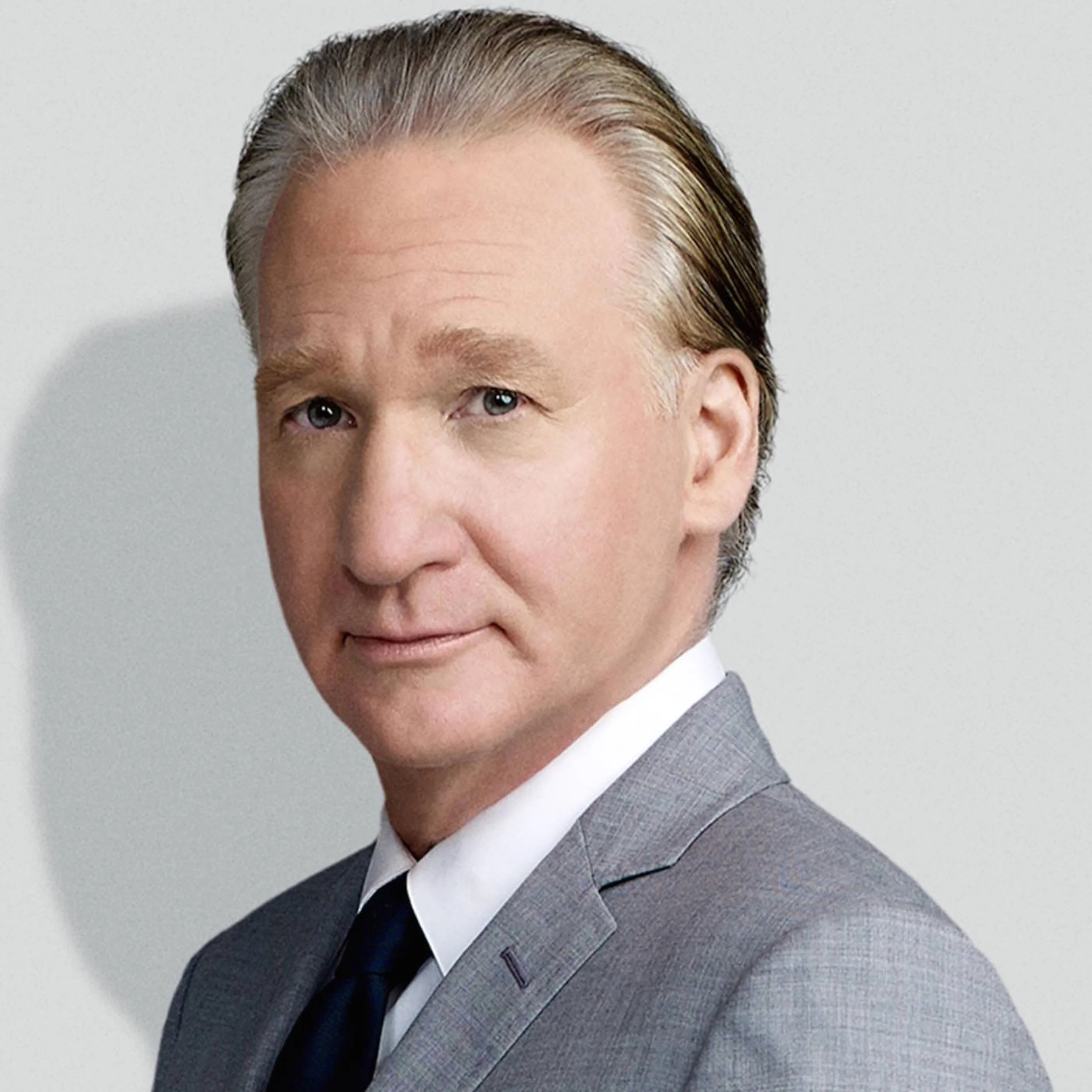Portrait of Bill Maher in a gray suit and dark tie against a light background