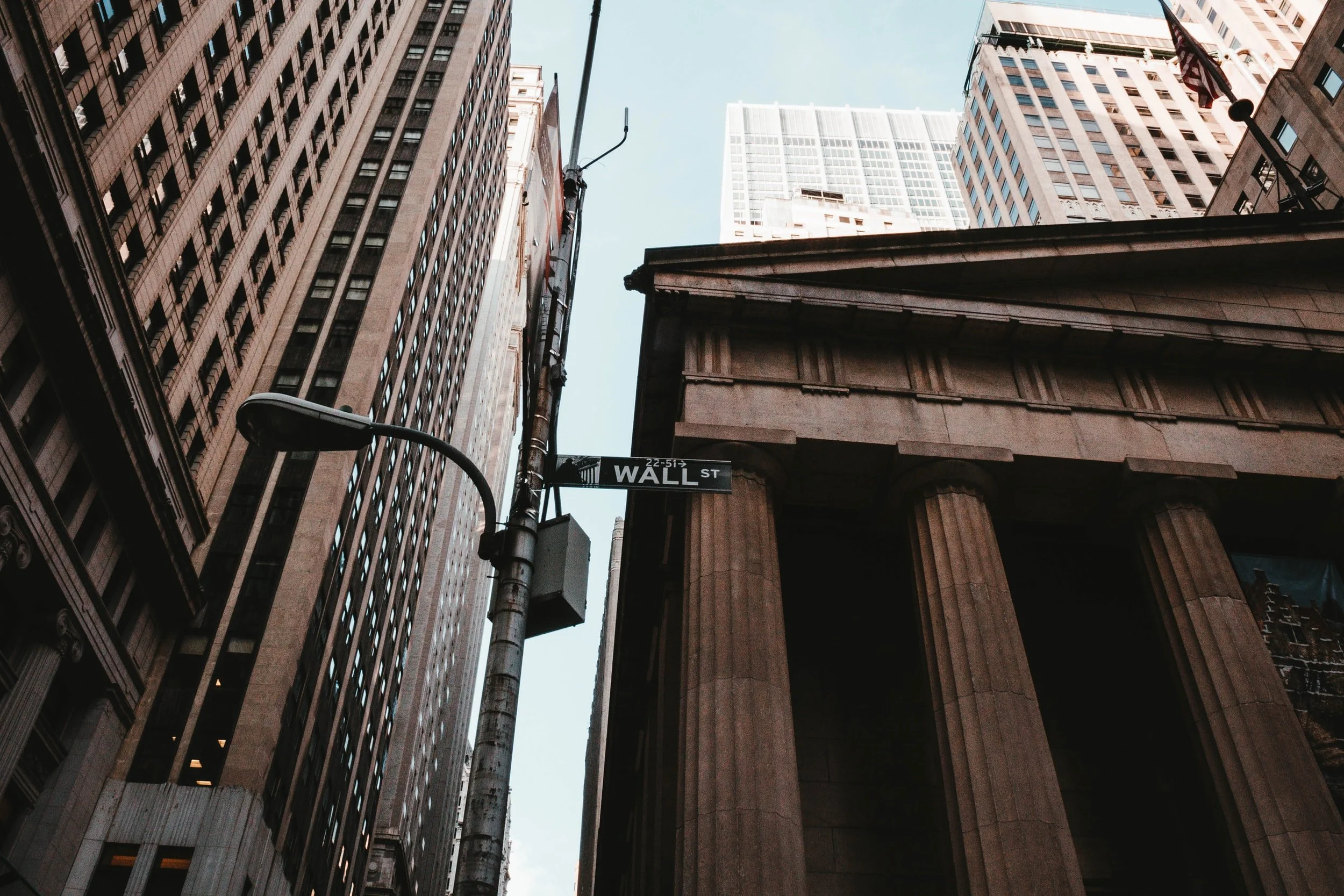 Looking up at a Wall St street sign between tall office towers and a building with stone columns