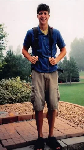 Tall smiling teenager in a blue polo, cargo shorts and backpack standing on a brick patio
