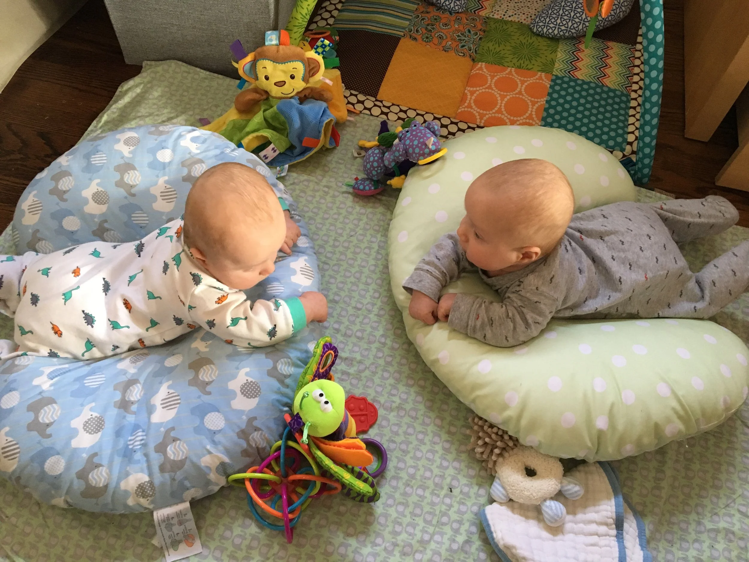 Twin babies doing tummy time on nursing pillows, facing each other among colorful toys
