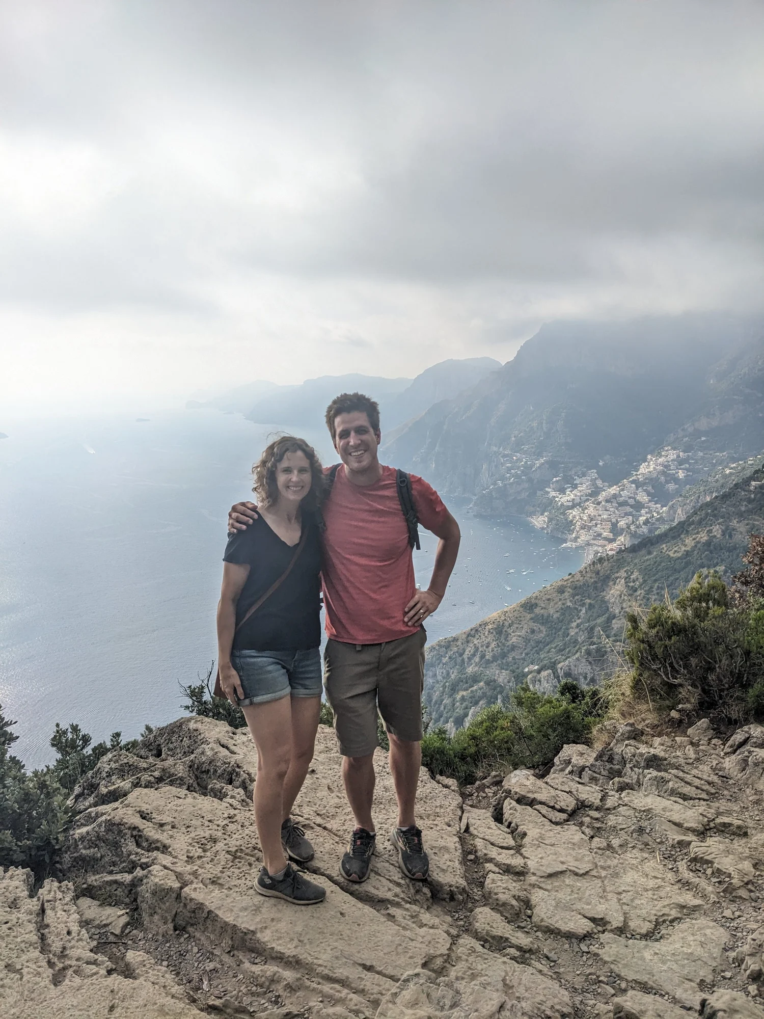 A smiling couple on a rocky cliff high above the sea and a hillside town on the Amalfi Coast