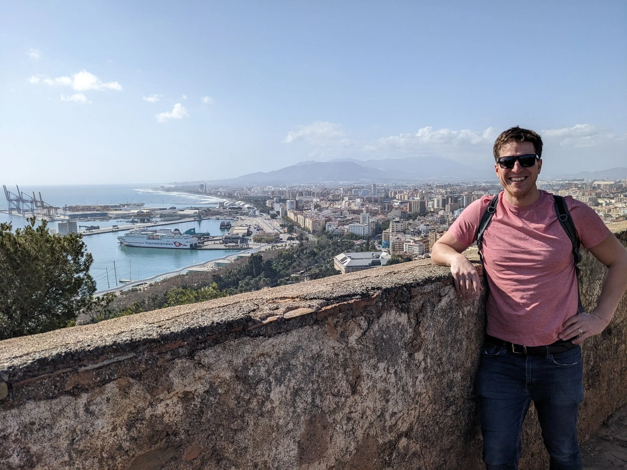 Smiling man in sunglasses and pink shirt leaning on a stone wall above Malaga's port and city