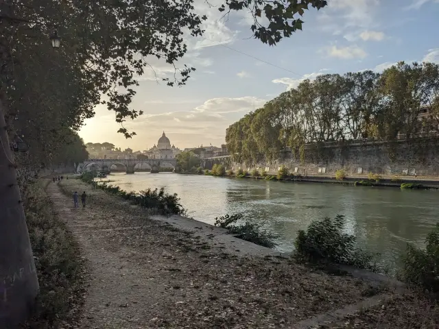 Tree-lined path along the Tiber River in Rome at sunset, with a stone bridge and St. Peter's dome in the distance