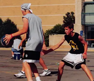 A young teen in a black shirt guards an older player dribbling a basketball on an outdoor court