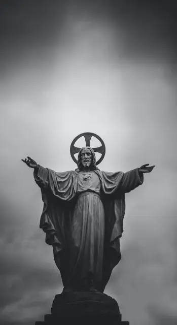 Black and white photo of a stone Jesus statue with arms outstretched and a halo, against a cloudy sky