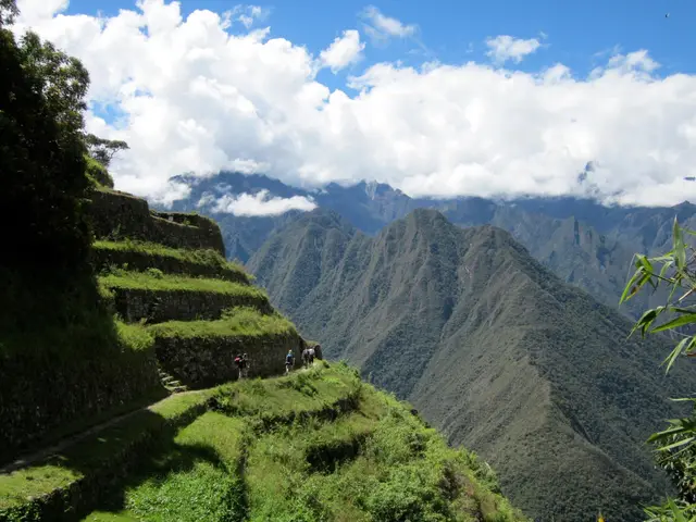 Hikers walking a trail past green stone terraces with steep Andes mountains and clouds behind