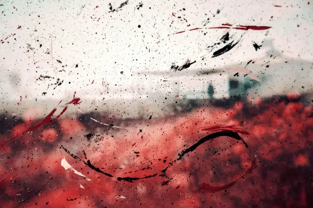 Black, red and white paint splattered across glass, with a blurred landscape and building behind it