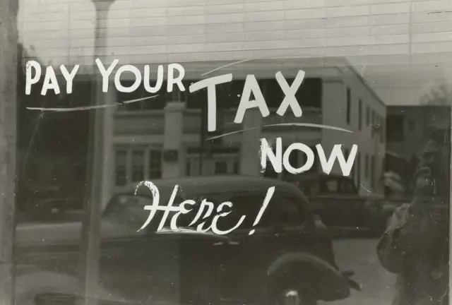 Old black-and-white photo of a shop window painted with "Pay your tax now here!" reflecting a street and vintage car.