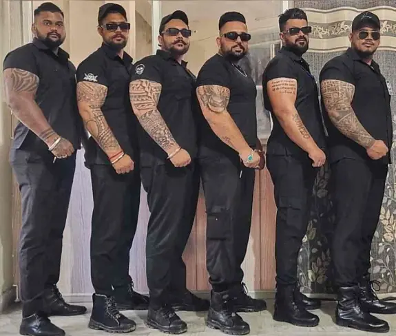 Six tattooed bouncers in black shirts, sunglasses and boots standing in a row