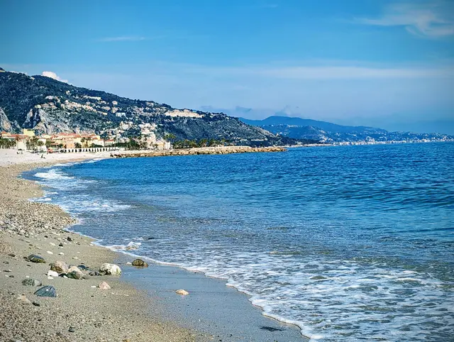 Pebbly Mediterranean beach with blue water curving toward a hillside town and distant coastline