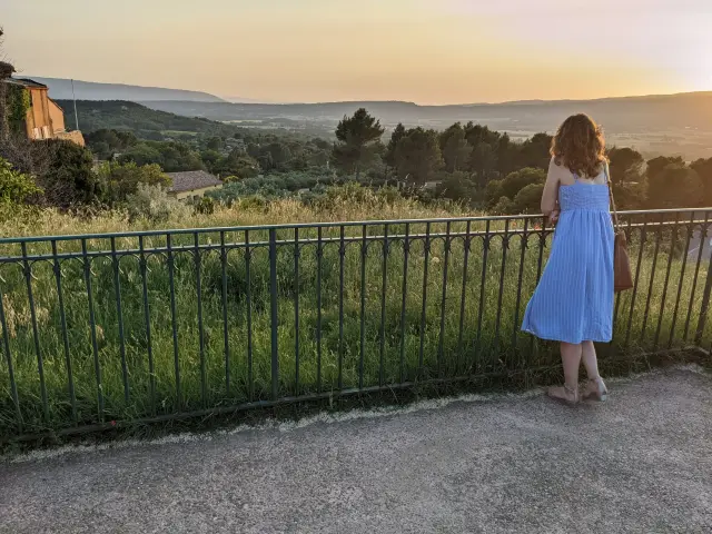 Woman in a blue sundress leaning on a railing, looking over a green Provence valley at sunset