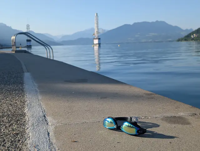 Mirrored swim goggles on a concrete lake edge with a ladder, diving tower and mountains in the background