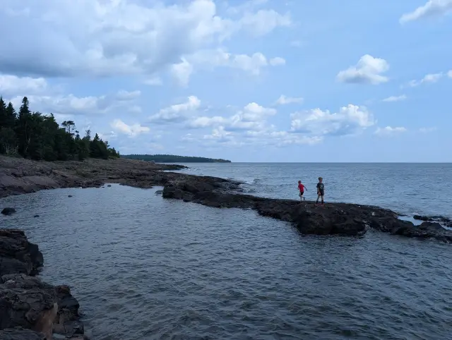 Two boys walking on a dark rocky point jutting into Lake Superior under a blue sky with clouds